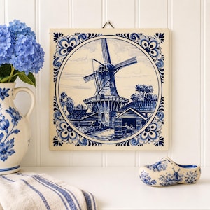 May include: A square ceramic wall tile featuring a blue and white illustration of a Dutch windmill within a circular frame. The tile is decorated with ornate floral corner patterns and is displayed alongside a matching blue and white ceramic clog.