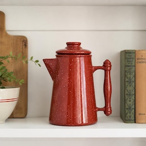 May include: A rust-red coffee percolator with a white speckled enamel finish, featuring a cylindrical body, a side handle, a spout, and a matching lid. The percolator is positioned centrally, flanked by a small potted plant and three vintage books.