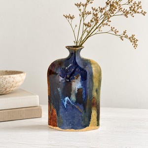 May include: A flat-shaped ceramic vase with a narrow neck and a glossy glaze featuring blended shades of deep blue, dark brown, and muted yellow. The vase has a rounded top and a flat base, containing a sprig of dried, light brown wildflowers.
