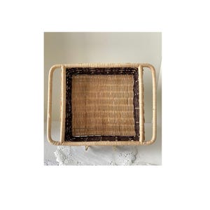 May include: Rectangular woven wicker tray with a dark brown border and handles. The tray has a natural tan color with a dark brown border and handles. The handles are curved and extend upwards from the sides of the tray.