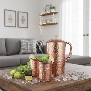 May include: A copper jug and two matching copper mugs on a wooden table with ice, lemons, and mint. The jug has a lid and handle. The mugs are filled with a dark liquid, garnished with lemon and mint. The table is 100 cm long.