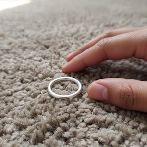 Könnte beinhalten: Ein einfacher, silberfarbener Ring. Der Ring ist kreisförmig und liegt auf einer strukturierten, hellbraunen Oberfläche. Der Ring scheint aus Metall zu sein und hat eine glatte, polierte Oberfläche. Der Ring ist ein minimalistisches Schmuckstück.