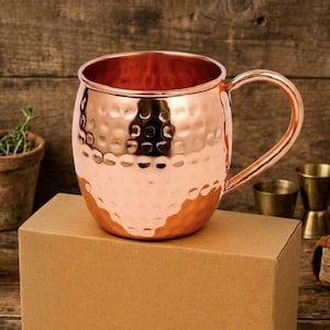 Puede incluir: Una taza de cerámica blanca con un asa negra y el texto "But First, Coffee" en un estilo manuscrito. La taza está sobre una superficie blanca. La taza mide aproximadamente 10 cm de alto.