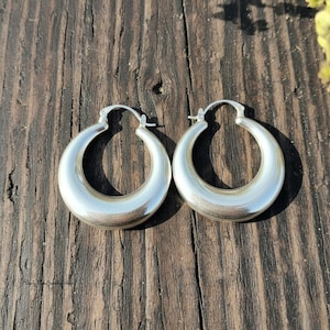 Puede incluir: Un par de pendientes de aro plateados con un diseño redondeado y grueso. Los pendientes tienen una superficie pulida y brillante. Se muestran sobre una superficie de madera marrón oscura y desgastada, con sombras.