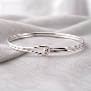 Könnte beinhalten: Ein silberfarbenes Armreif-Armband mit einem tropfenförmigen Verschluss und einem kleinen, kugelförmigen Verschluss. Das Armband hat ein schlichtes, elegantes Design mit drei parallelen Bändern. Der Hintergrund ist ein weicher, grauer Stoff.