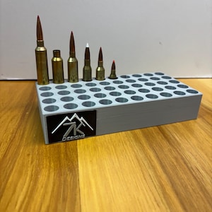 May include: A gray ammunition storage tray with multiple holes, holding various sizes of brass and copper bullets with red tips. The tray has a black and white logo that says "7K DESIGNS".