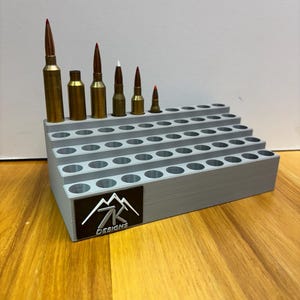 May include: A gray, tiered ammunition holder with multiple circular openings, holding various sizes of brass and copper-colored bullets with red and white tips. The holder has a black label with a white mountain and the text "7K Designs".