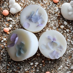 Op de afbeelding: Drie ronde, witte wassmeltkernen met ingebedde blauwe en paarse bloemblaadjes. De smeltkernen liggen op een getextureerd, grijs oppervlak met kleine kiezels en verspreide decoratieve elementen.