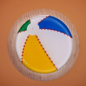 May include: A round, colorful beach ball design on a wooden coaster. The ball features segments of white, blue, yellow, and green, with red stitching details. The coaster is made of light-colored wood.