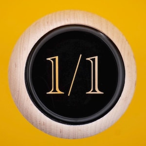 Może przedstawiać: Okrągły obiekt z tekstem "1/1" w kroju szeryfowym, na czarnym tle. Czarny okrąg jest obramowany jasnym drewnianym pierścieniem, wszystko na jasnym żółtym tle.