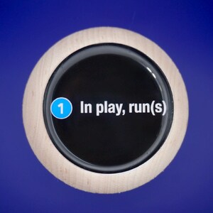 Może przedstawiać: Okrągły przedmiot z drewnianą ramą i czarnym środkiem. Tekst "1 In play, run(s)" jest wyświetlany na biało. Niebieskie kółko z numerem "1" znajduje się po lewej stronie. Tło jest jednolicie niebieskie.