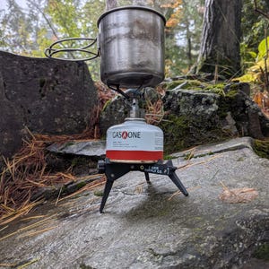 Puede incluir: Una olla de camping de acero inoxidable con un asa de alambre se encuentra sobre una estufa de camping portátil. La estufa está conectada a un cartucho de combustible Gas One. Sale vapor de la olla.