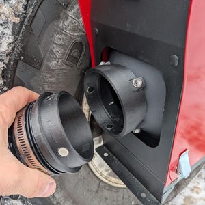 May include: Black metal exhaust component with a clamp, positioned near a red panel and a black tire. The component is being held near a matching opening on the red panel. The tire has a deep tread pattern.
