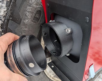 Op de afbeelding: Zwart metalen uitlaatcomponent met een klem, in de buurt van een rood paneel en een zwarte band. Het component wordt in de buurt van een bijpassende opening op het rode paneel gehouden. De band heeft een diep profiel.