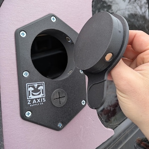 May include: Black, hexagonal electrical outlet cover with a hinged lid. The cover is mounted with screws and features the text "Z AXIS SUPPLY CO." and a plus sign. The lid has a magnetic closure.