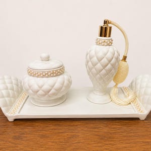 May include: A white ceramic vanity set featuring a rectangular tray with quilted edges, a lidded jar with a quilted pattern and pearl-like trim, and a perfume bottle with a gold pump, quilted body, pearl-like collar, and a cream-colored tassel.