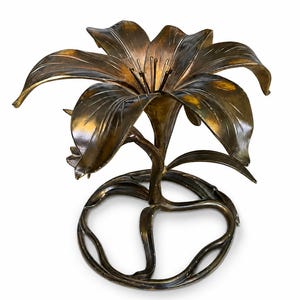 May include: A bronze-toned metal sculpture shaped like a lily flower. The piece features a large, open bloom with curved petals and a central stamen, supported by a stem that extends downward into a circular, vine-like base structure.