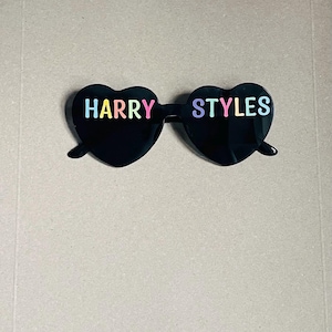 May include: Black heart-shaped sunglasses with the words "HARRY STYLES" in colourful letters. The sunglasses have a classic frame and are set against a neutral background.