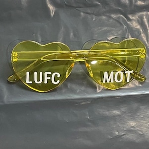 Puede incluir: Gafas de sol amarillas en forma de corazón con las inscripciones blancas «LUFC» y «MOT». Las gafas tienen montura y lentes transparentes. La imagen está tomada sobre una superficie oscura y texturizada.