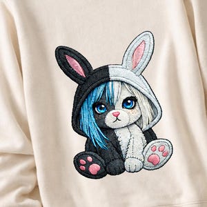 Könnte beinhalten: Cremefarbenes Sweatshirt mit einem gestickten Design eines Hasen-Katzen-Hybriden. Das Design ist in der Mitte geteilt, eine Seite schwarz und die andere weiß. Die Hasenohren und Pfoten haben rosa Akzente, und das Tier hat blaue Augen.
