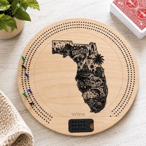 May include: A round wooden cribbage board featuring a laser-engraved map of Florida filled with detailed illustrations of landmarks and wildlife. The board includes a circular track of holes for scoring, colored pegs, and the word Wins printed below.