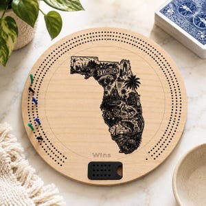 May include: A round wooden cribbage board featuring a laser-engraved map of Florida filled with detailed illustrations of local landmarks, wildlife, and the word FLORIDA. The board includes scoring holes, colored pegs, and the word Wins near a black slot.