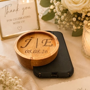 May include: A wooden wedding ring holder with the initials "J | E" and the date "08.08.26" engraved on it. The holder sits on a black phone case. A sign reads "Thank you for celebrating with us!"