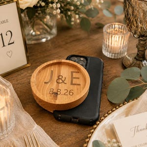 May include: A round wooden coaster featuring the engraved letters J & E and the date 8.8.26, resting on top of a black smartphone. The coaster has a raised circular rim and a natural wood grain finish, positioned on a table setting.