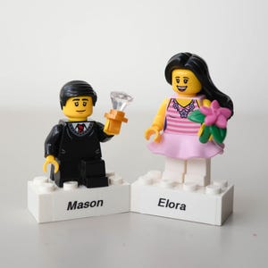 May include: Two Lego figurines, a man in a black suit holding a diamond ring and a woman in a pink dress holding flowers. Each figure stands on a white base with a name: "Mason" and "Elora".