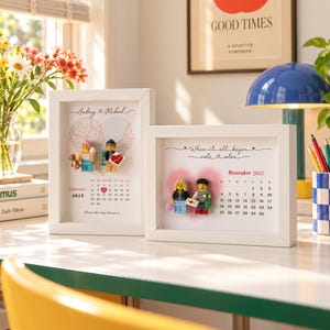 May include: Two white framed shadow boxes featuring personalized calendars and miniature plastic figures. The left frame displays September 2023 with Audrey and Michael, while the right frame shows November 2025 with Cole and Alex, both with heart motifs.