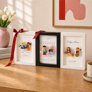 Puede incluir: Tres marcos de caja rectangulares que contienen minifiguras personalizadas y texto impreso. Dos marcos son blancos con cintas rojas y uno es negro. Las pantallas muestran nombres personalizados, fechas y pequeñas figuras de plástico con accesorios.