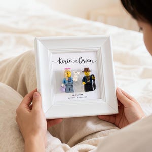 May include: A white framed artwork with Lego figures depicting a couple and a small dog. The names "Korie & Brian" are written above the figures, with the date "14.06.2024" and the phrase "ALWAYS & FOREVER" below.