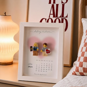 May include: A white shadow box frame containing a personalized calendar print for July 2024. The print features the names destiny and steven, a pink heart graphic, and two plastic minifigures. One figure holds a chocolate bar, the other a heart card.