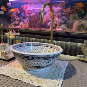 May include: A decorative white and blue patterned ceramic sink bowl with a brass faucet. The sink is placed on a white lace doily on a woven table. The background features an aquarium with fish.
