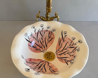 May include: A round, cream-colored ceramic vessel sink with a scalloped edge, featuring hand-painted black floral line art over soft pink circular brushstrokes. A brass-toned metal faucet is positioned directly above the basin.