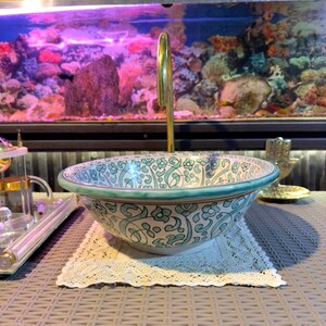 May include: A decorative ceramic sink with a floral pattern in teal and white. The sink has a rounded shape and sits on a cream-colored lace doily. A gold-colored faucet rises from the center.