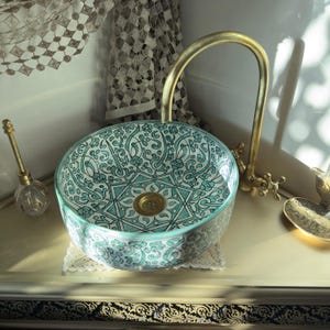 May include: A round ceramic sink with a teal and white floral pattern. The sink has a gold-colored faucet and drain. The sink is on a cream-colored surface with a decorative lace doily. The overall theme is bathroom decor.