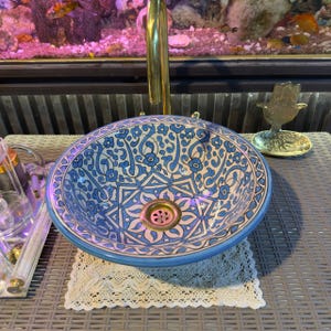 May include: A round, ceramic sink with a blue and white floral pattern. The sink has a brass drain and is set on a gray surface. A gold faucet is above the sink. A small gold hand-shaped object is to the right of the sink.