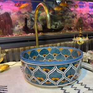 May include: A decorative ceramic sink with a blue, teal, and yellow floral pattern. The sink is round and sits on a white surface. A gold-colored faucet is positioned above the sink. The background includes an aquarium with fish.