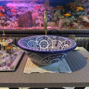 May include: A decorative ceramic sink with a blue and white pattern. The sink has a rounded shape with a wide rim and a gold-colored faucet. The design features intricate geometric and floral motifs, creating an elegant and artistic appearance. The sink is placed on a gray table.
