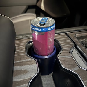 Könnte beinhalten: Eine rosa-silberne Dose zuckerfreies Energy-Drink in einem dunkelblauen Getränkehalter. Die Dose befindet sich in der Mittelkonsole eines Autos mit Holzmaserung. Auf der Dose steht "SUGARFREE".