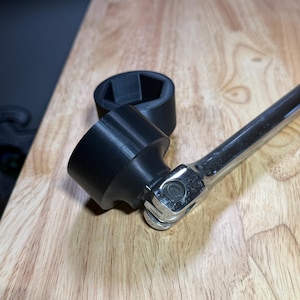 May include: A black, 3D-printed socket adapter connected to a chrome ratchet wrench. The adapter features a hexagonal opening. The items are on a light brown wooden surface.