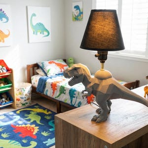 May include: A child's bedroom with a dinosaur theme. The room features a bed with a dinosaur-patterned comforter, a rug with dinosaur designs, and artwork on the wall. A lamp with a dinosaur-shaped base sits on a wooden table. Books and toys are also visible.