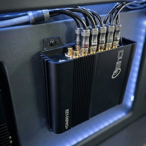 May include: Black car audio amplifier with multiple input and output connectors. The amplifier is mounted on a black panel and has the text "EZY-DSP 612+" and "DLS" printed on it. Several black cables are connected to the amplifier.