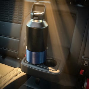 Puede incluir: Una botella de agua negra aislada con borde y asa plateados, colocada en un portavasos azul. La botella está en la consola central de un coche, con luz solar.