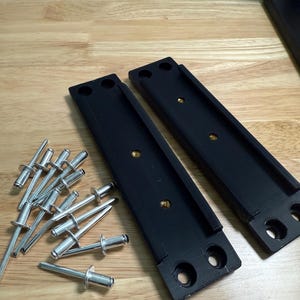 May include: Two black rectangular brackets with multiple holes and brass inserts, alongside a collection of silver rivets. The items are arranged on a light brown wooden surface, suggesting a hardware or construction theme.