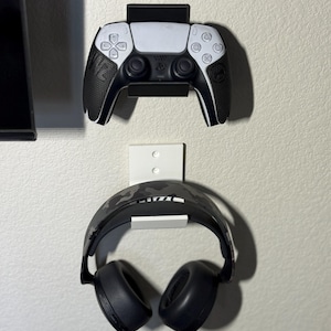 May include: A black and white game controller and a pair of black headphones mounted on a wall. The controller has a white center and black sides. The headphones have a camouflage pattern on the headband.