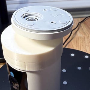 May include: A white and light gray security camera with a cylindrical body and a circular top. The top has the word "UP" and arrows. The camera is on a black surface with white dots.