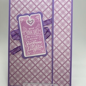 Puede incluir: Una tarjeta de felicitación con un patrón de cuadros morados y blancos. Una etiqueta morada con las palabras "May Your Day Be Filled With Sunshine & Laughter" está sujeta con una cinta morada. La tarjeta está sostenida por un soporte de plástico transparente.