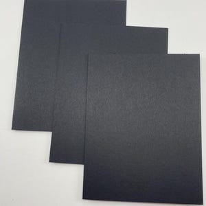 Pode incluir: Uma pilha de tr&ecirc;s folhas de papel cart&atilde;o preto. O papel tem uma superf&iacute;cie ligeiramente texturizada e &eacute; retangular. As folhas est&atilde;o dispostas em um padr&atilde;o escalonado, com cada folha sobrepondo-se parcialmente &agrave; de baixo.